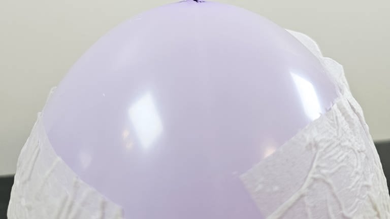 Wasser auf den Boden des Luftballons tropfen oder sprühen und ein Blatt Klopapier auf die feuchte Stelle legen. Nochmals Wasser auf das Papier geben bis alle Stellen möglichst flach und glatt am Luftballon anliegen.