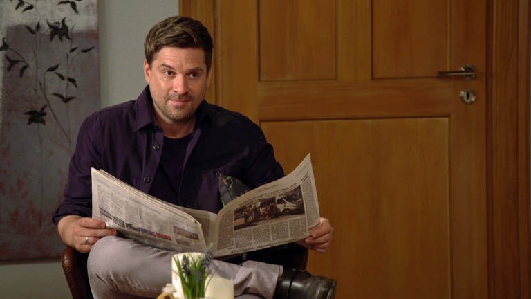 Andreas sitzt im Wohnzimmer und liest Zeitung.