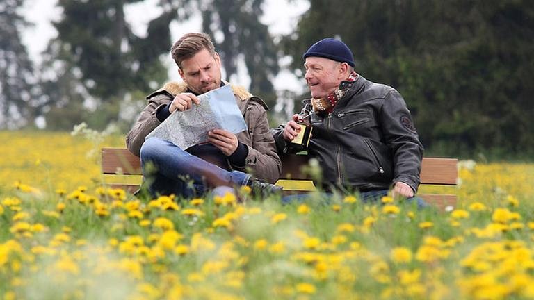 Andreas und Toni sitzen auf einer Bank, Toni hält ein Bier, Andreas studiert eine Landkarte, ringsum blüht der Löwenzahn