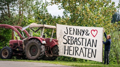 Sebastian hält ein riesiges Stück Stoff, das an einem Traktor befestigt ist. Darauf steht "Jenny und Sebastian heiraten" (Foto: SWR, d:light/Christian Koch)