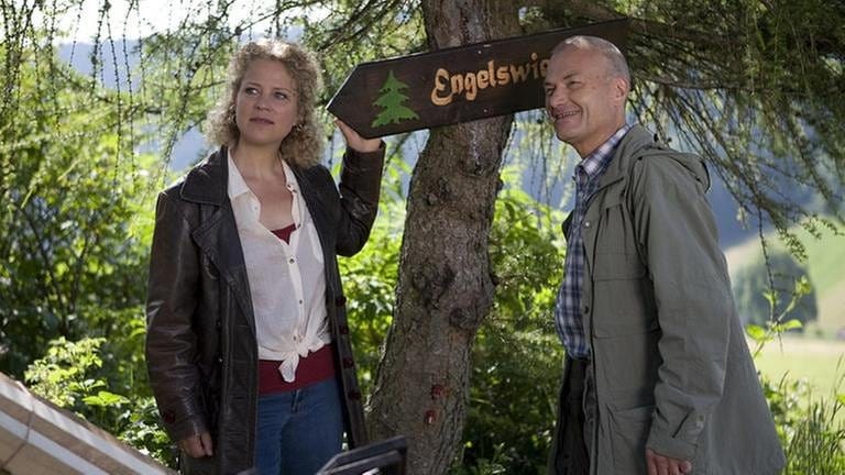 Bea und Karl stehen an einem Baum, an dem sie das Hinweisschild zur Engelswiese angebracht haben