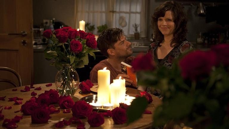 Andreas kniet vor Eva, die am Tisch sitzt, der mit Kerzen, einem Strauß roter Rosen und Rosenblättern geschmückt ist