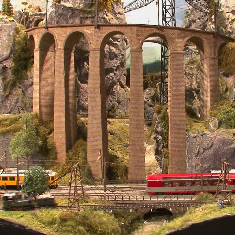 Ein gelber und ein roter Zug einer Modelleisenbahn mit Felsen und Brücke