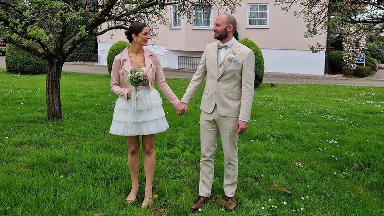 Paar im Garten zeigt Brautkleid und Anzug für die Hochzeit