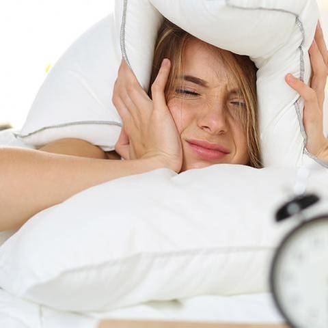 Eine Frau liegt im Bett und hält sich mit müdem Blick auf einen Wecker das Kopfkissen über die Ohren.