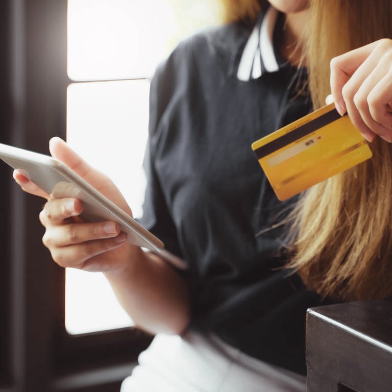 Frau mit langen dunkelblonden Haaren lehnt sich an eine Theke - in der einen Hand ihr Smartphone und in der anderen eine gelbe Kreditkarte.