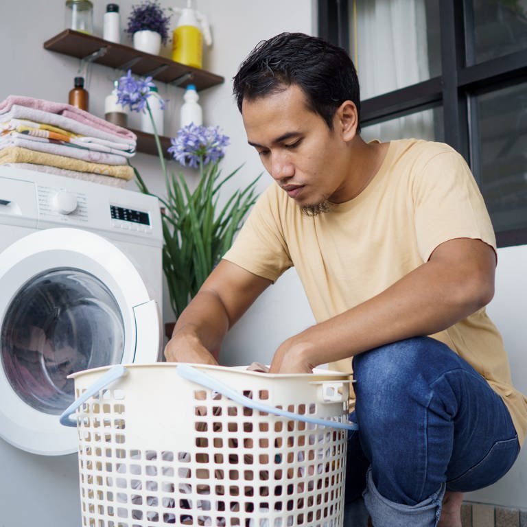 Junger Mann befüllt eine Waschmaschine mit Wäsche. Was sagt die Füllmenge bei der Waschmaschine aus? Welche ist wirklich effizient?