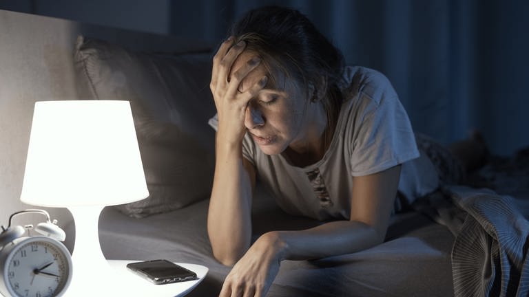Frau liegt auf dem Bauch im Bett und hält sich schmerzverzerrt das Gesicht. Chronische Schmerzen: Wie entsteht ein Schmerzgedächtnis? Welche Therapie hilft?