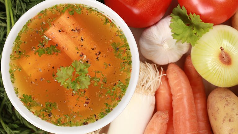 Eine Schale klare Gemüsebrühe und die Zutaten für den Fond. Klare Brühe aus Rind, Huhn, Fisch oder Gemüse ist die ideale geschmackliche Grundlage für Suppen.
